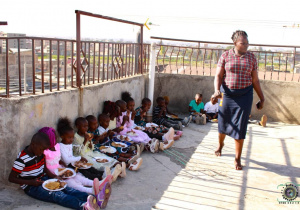 Zdjęcie przedstawia murek pod którym siedzą kenijskie dzieci i jedzą posiłek.